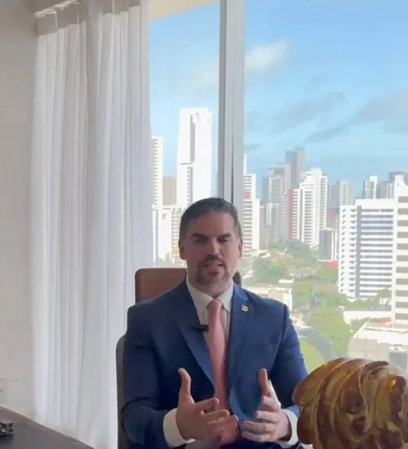 Dr. Guilhermino sentado à mesa da sala de consulta, com a cidade ao fundo